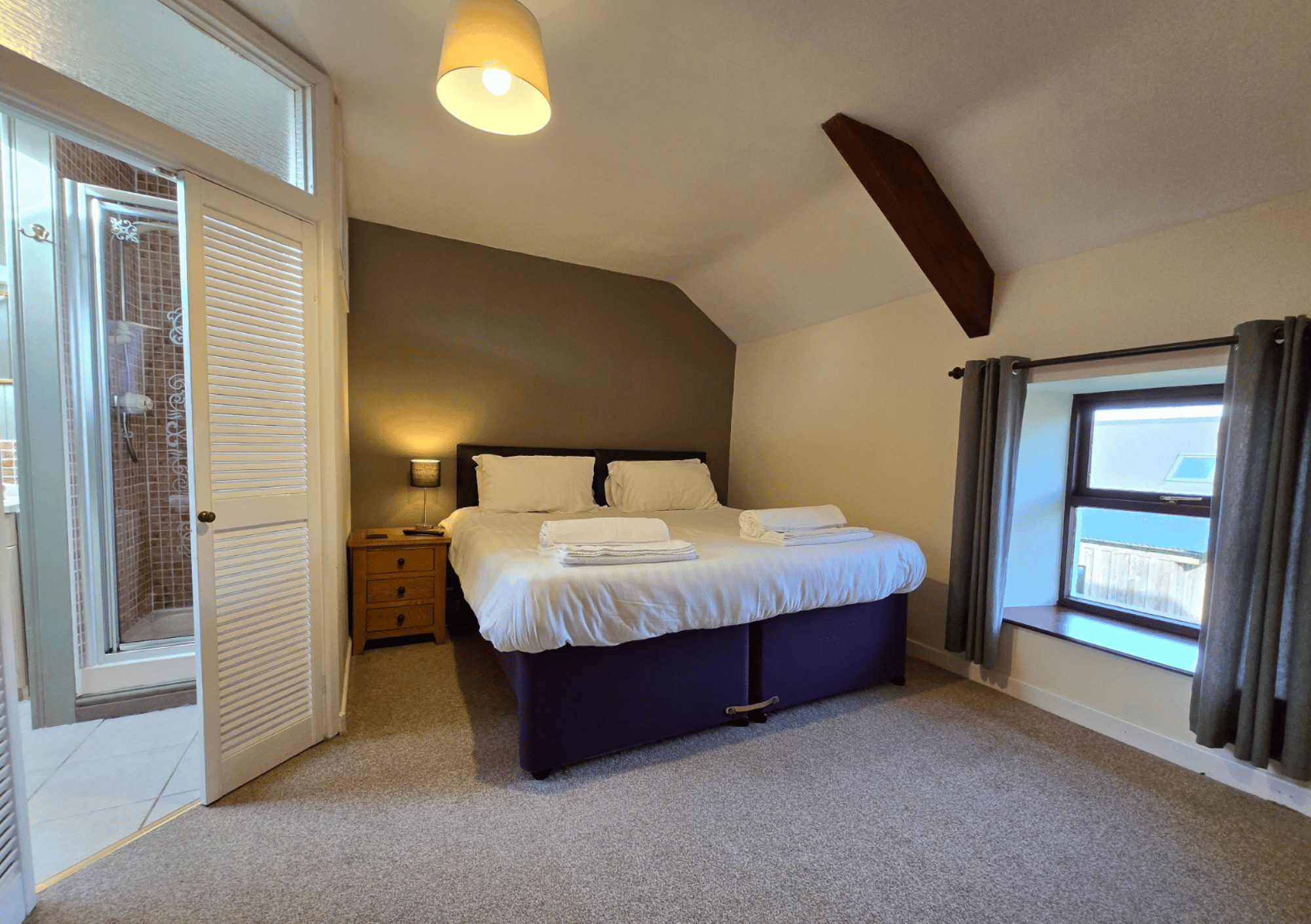 Hotel bedroom with a king bed, white linens, two pillows, a wooden nightstand, and a slanted ceiling beam above the headboard.